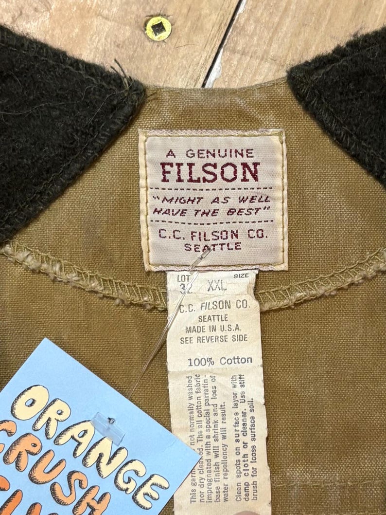 Pu&ograve; includere: Primo piano di un'etichetta di giacca Filson beige con il testo "A Genuine Filson" e "Might as well have the best". L'etichetta include anche "C.C. Filson Co. Seattle" e "Made in U.S.A." Un'etichetta con "Orange Crush" &egrave; visibile.