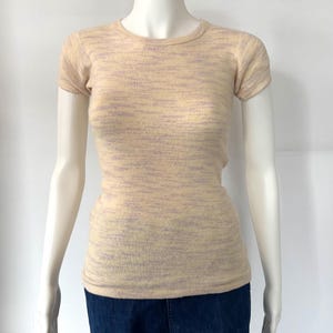 Könnte beinhalten: Ein kurzärmeliges T-Shirt mit Rundhalsausschnitt in hellem Beige mit einem dezenten, strukturierten Muster aus Lavendel und Beige. Das Shirt wird auf einer Schaufensterpuppe präsentiert, kombiniert mit dunkelblauen Jeans.