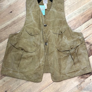 Pu&ograve; includere: Un gilet in cotone cerato color beige con pi&ugrave; tasche e chiusura con bottoni. Il gilet ha un colletto nero e un'etichetta con testo. Progettato per attivit&agrave; all'aperto.