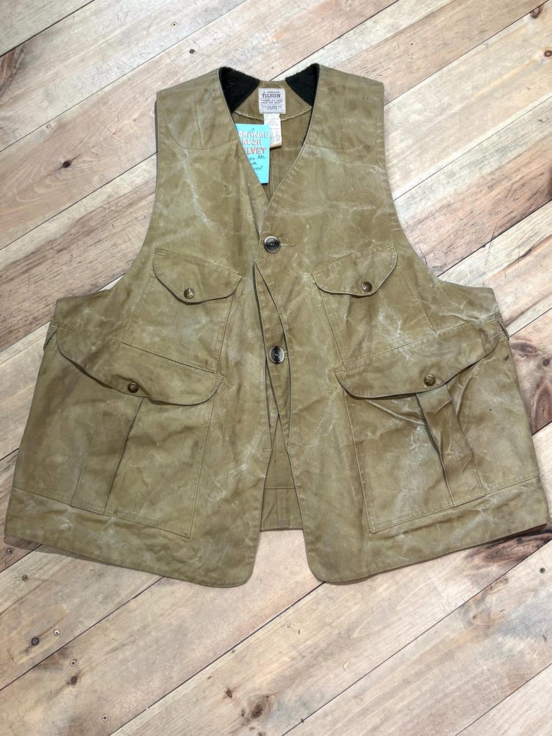 Pu&ograve; includere: Un gilet in cotone cerato color beige con pi&ugrave; tasche e chiusura con bottoni. Il gilet ha un colletto nero e un'etichetta con testo. Progettato per attivit&agrave; all'aperto.