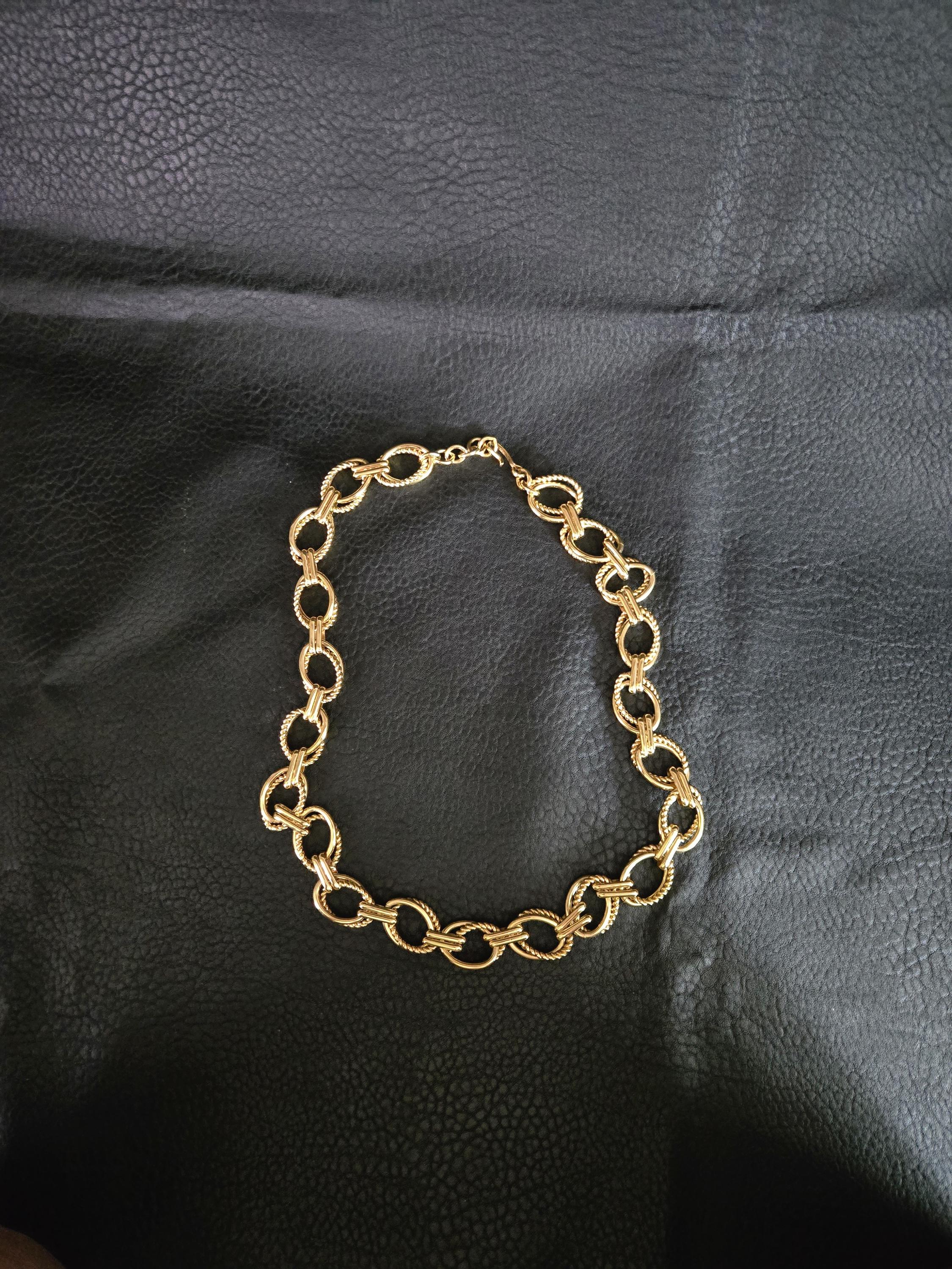 May include: A gold-colored necklace with a chain link design. The necklace is made of oval-shaped links, with a textured pattern on the surface. The necklace is displayed on a black surface.