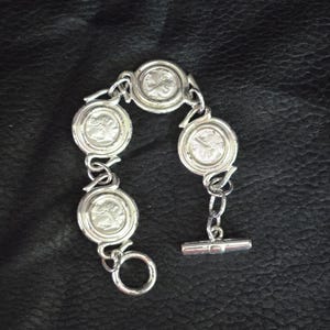 May include: Silver-toned bracelet featuring five circular medallions with a raised design. The bracelet has a toggle clasp and is linked with small, curved connectors. The jewelry is displayed on a dark, textured surface.