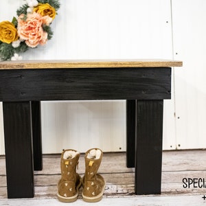 May include: A black wooden bench with a gold top. The bench has four legs and is sitting on a wooden floor. There are two pairs of brown boots with white polka dots sitting in front of the bench. The text "SPECIAL WALNUT + BLACK" is written on the floor in front of the bench.