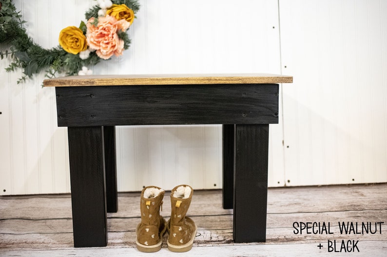 May include: A black wooden bench with a gold top. The bench has four legs and is sitting on a wooden floor. There are two pairs of brown boots with white polka dots sitting in front of the bench. The text "SPECIAL WALNUT + BLACK" is written on the floor in front of the bench.