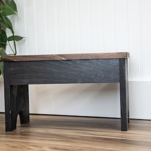 May include: A black wooden stool with a dark brown top. The stool has a simple design with straight legs and a rectangular top.