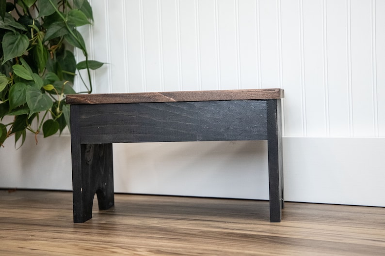 May include: A black wooden stool with a dark brown top. The stool has a simple design with straight legs and a rectangular top.