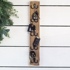 May include: A wooden key holder with five hooks, each holding a set of keys. The key holder is mounted on a white wall.