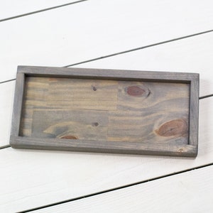 Op de afbeelding: Een rechthoekige houten dienblad met een grijze beitsafwerking. Het dienblad heeft een verhoogde rand en is gemaakt van natuurlijk hout met knoesten en nerven.