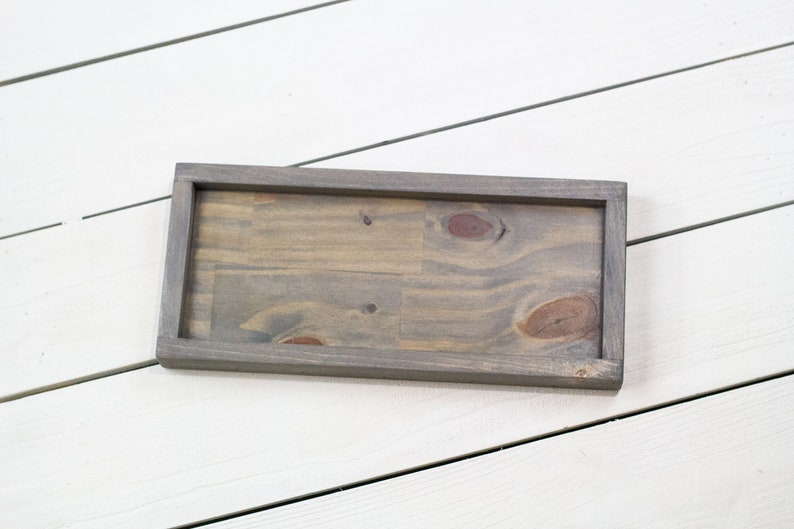 Op de afbeelding: Een rechthoekige houten dienblad met een grijze beitsafwerking. Het dienblad heeft een verhoogde rand en is gemaakt van natuurlijk hout met knoesten en nerven.
