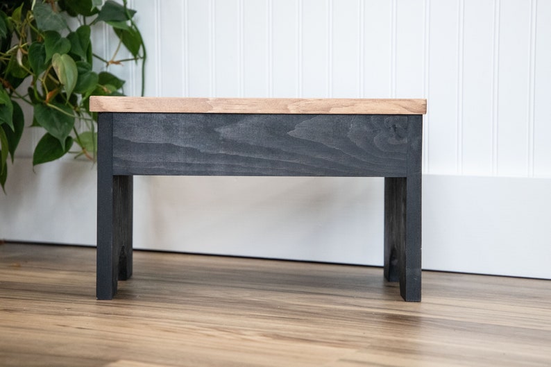 May include: A small wooden stool with a black stained top and legs. The stool is made of wood and has a simple design.