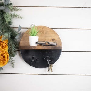 May include: A round wooden key holder with a black shelf and two hooks. The key holder has a natural wood finish on the top half and a black painted finish on the bottom half. There is a small potted plant and a pair of glasses on the shelf.