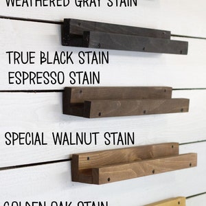 Op de afbeelding: Vijf houten planken in verschillende beitskleuren, gemonteerd aan een witte muur. De planken zijn gelabeld met hun beitskleuren: Weathered Gray Stain, True Black Stain, Espresso Stain, Special Walnut Stain en Golden Oak Stain.
