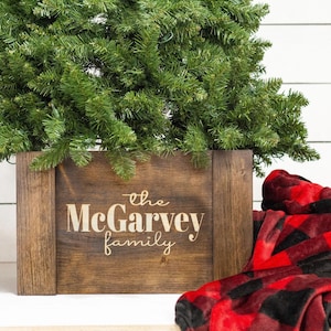 May include: A wooden Christmas tree stand with the text "the McGarvey family" engraved on the front. A red and black plaid blanket is in the foreground.