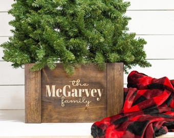 May include: A wooden Christmas tree stand with the text "the McGarvey family" engraved on the front. A red and black plaid blanket is in the foreground.