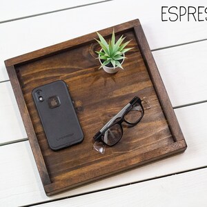 Op de afbeelding: Een houten dienblad met een donkerbruine vlek, met een zwarte telefoon in een hoesje, een paar zwarte brillen en een kleine groene plant in een pot. Het dienblad is gelabeld met "ESPRESSO".