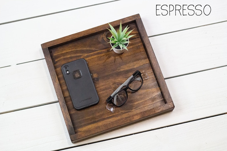Op de afbeelding: Een houten dienblad met een donkerbruine vlek, met een zwarte telefoon in een hoesje, een paar zwarte brillen en een kleine groene plant in een pot. Het dienblad is gelabeld met "ESPRESSO".