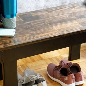 May include: A dark brown wooden bench with black legs. The bench is sitting on a light wood floor. There are two pairs of shoes under the bench, one pair is pink and the other is gray.