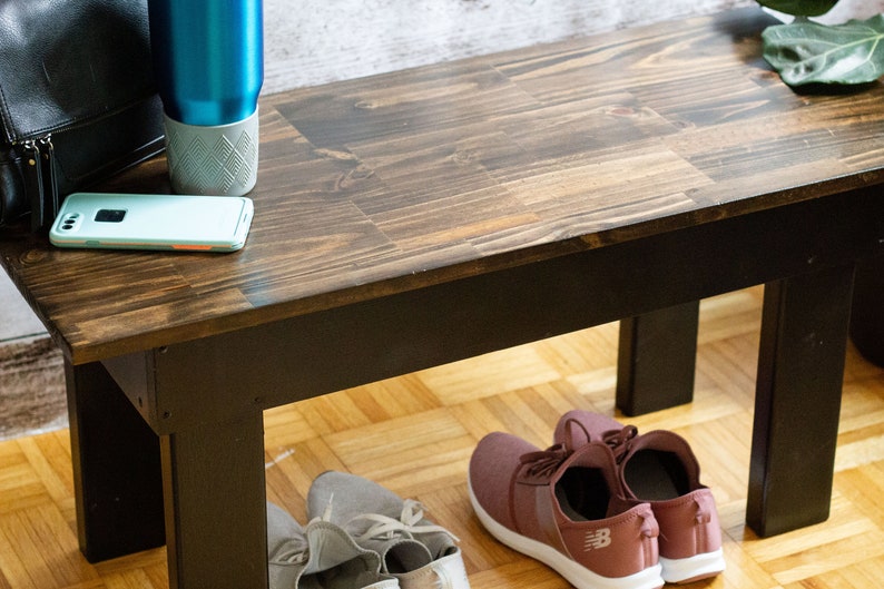 May include: A dark brown wooden bench with black legs. The bench is sitting on a light wood floor. There are two pairs of shoes under the bench, one pair is pink and the other is gray.