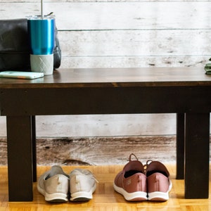 May include: A dark brown wooden bench with black legs. Two pairs of shoes are placed under the bench. One pair is light grey and the other is pink.