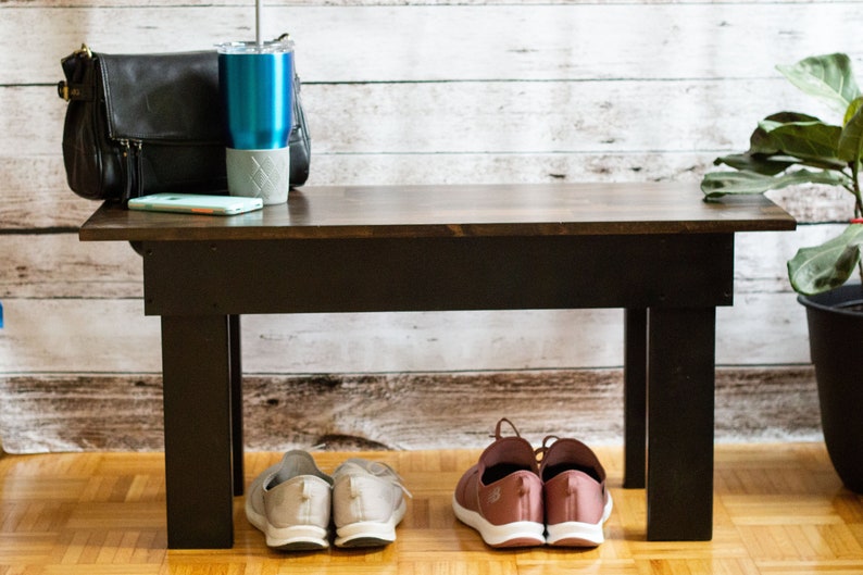 May include: A dark brown wooden bench with black legs. Two pairs of shoes are placed under the bench. One pair is light grey and the other is pink.