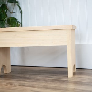 May include: A simple, unfinished wooden stool with a rectangular top and two legs on each side. The legs have a slight curve at the bottom.