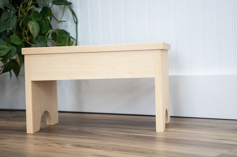 May include: A simple, unfinished wooden stool with a rectangular top and two legs on each side. The legs have a slight curve at the bottom.