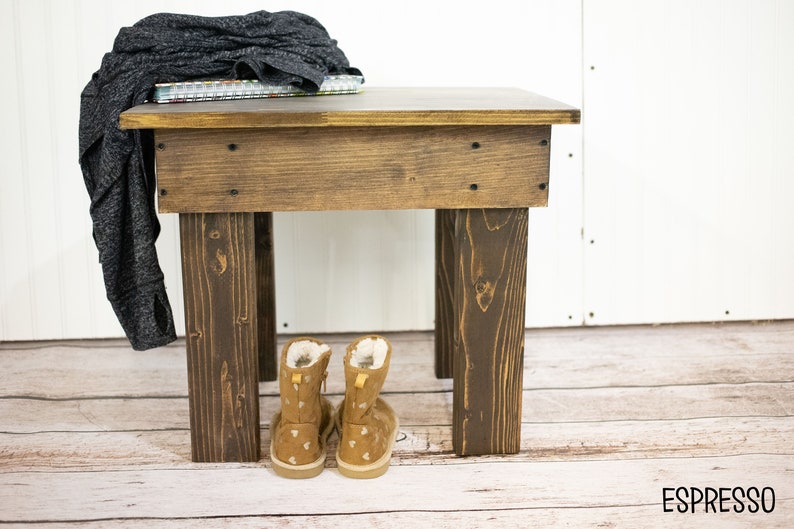 May include: A dark brown wooden end table with a square top and four legs. A gray sweater and a stack of books are on top of the table. Two brown suede boots with white accents are under the table. The text "ESPRESSO" is on the floor.