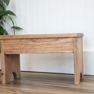 May include: A wooden stool with a natural brown finish. The stool has a simple design with a rectangular top and two legs on each side.