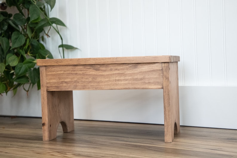 May include: A wooden stool with a natural brown finish. The stool has a simple design with a rectangular top and two legs on each side.