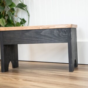 May include: A small wooden stool with a black stained base and a natural wood top. The stool has a simple design with rounded legs.