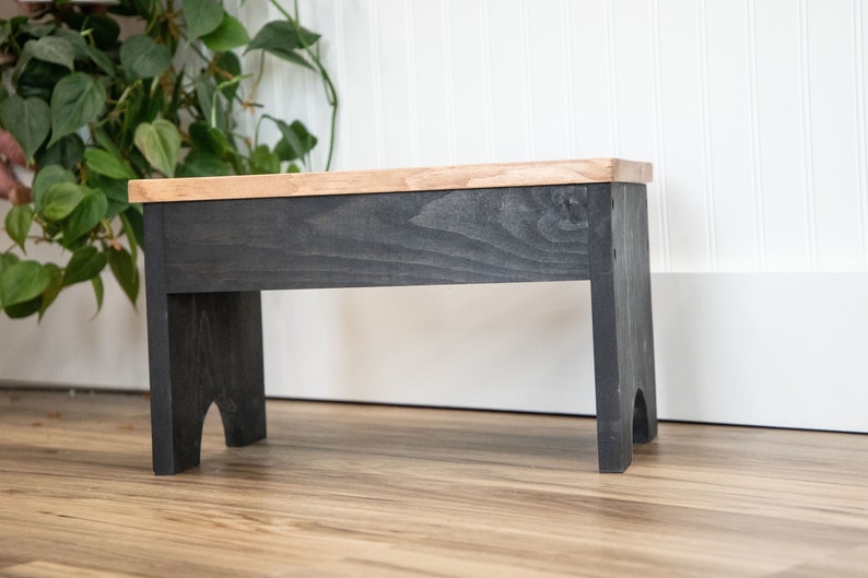 May include: A small wooden stool with a black stained base and a natural wood top. The stool has a simple design with rounded legs.
