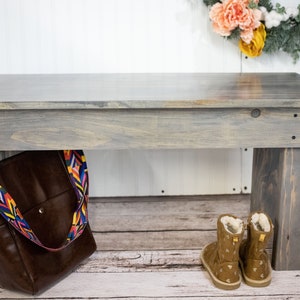 May include: A gray wooden bench with a brown leather bag hanging from the side. The bench has a distressed finish and is made of reclaimed wood. The bag has a colorful strap with a geometric pattern. The bench is sitting on a white wooden floor.
