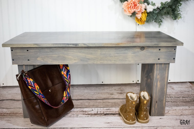 May include: A gray wooden bench with a brown leather bag hanging from the side. The bench has a distressed finish and is made of reclaimed wood. The bag has a colorful strap with a geometric pattern. The bench is sitting on a white wooden floor.