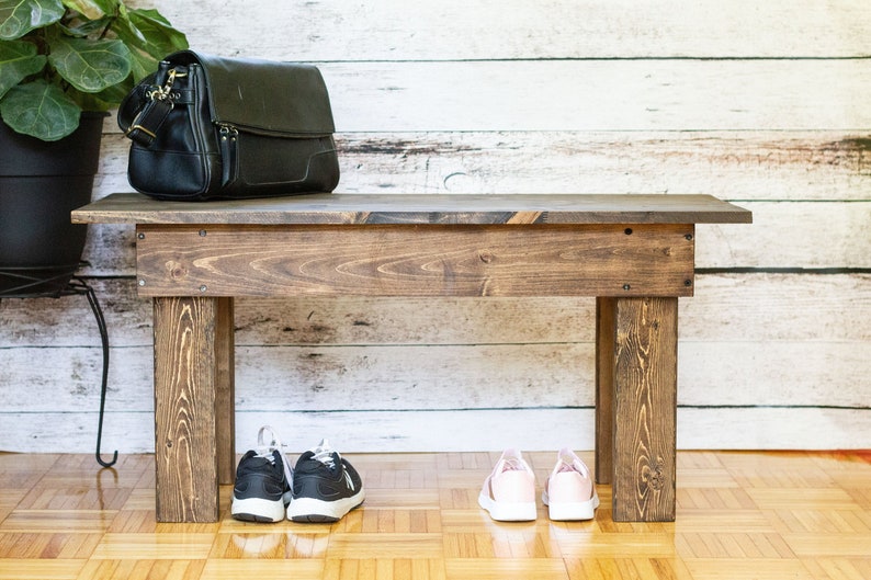 May include: A wooden bench with a dark brown stain. A black leather bag is sitting on top of the bench. There are two pairs of shoes on the floor in front of the bench, one pair is black and the other is pink.