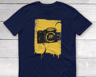 May include: A navy blue t-shirt with a yellow graphic of a Pentax camera with dripping paint.