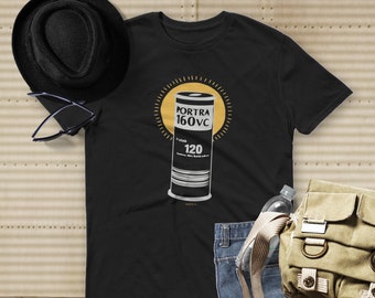 May include: Black t-shirt featuring a graphic of a Kodak Portra 160VC film roll inside a yellow sunburst design. The shirt is accompanied by a black hat, sunglasses, blue jeans, a water bottle, and a canvas bag. The text "120" is visible on the film roll.