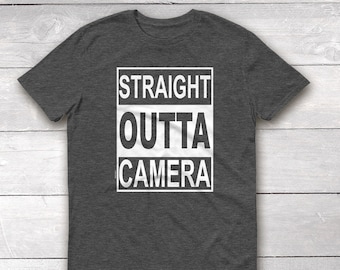 May include: A dark heather grey t-shirt with a white graphic that says "STRAIGHT OUTTA CAMERA" in a box.