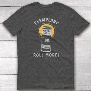 May include: A dark gray t-shirt with the text "EXEMPLARY" above a graphic of a roll of Portra 160VC film and the text "ROLL MODEL" below the graphic.