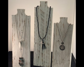May include: Three whitewashed wooden necklace displays with different necklaces. The display on the left has a layered necklace with a turquoise pendant. The middle display has a black beaded necklace with a silver cross pendant. The display on the right has a silver chain necklace with a silver pendant.