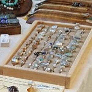 May include: A wooden display tray filled with a variety of silver and gold rings with different gemstones. The rings are arranged in rows on a beige fabric lining.