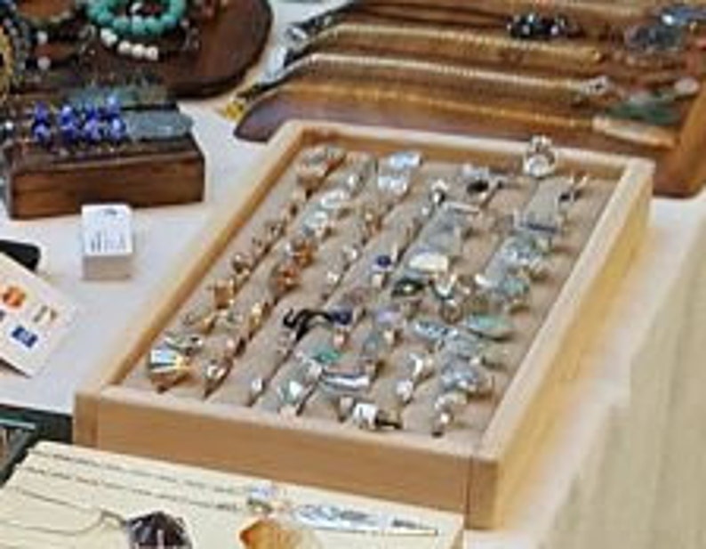 May include: A wooden display tray filled with a variety of silver and gold rings with different gemstones. The rings are arranged in rows on a beige fabric lining.