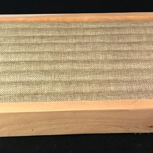 May include: A wooden ring display box with a light brown finish. The box has a fabric lining with a light brown color and a textured pattern. The box is designed to hold and display rings.