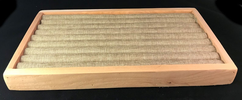 May include: A wooden ring display box with a light brown finish. The box has a fabric lining with a light brown color and a textured pattern. The box is designed to hold and display rings.