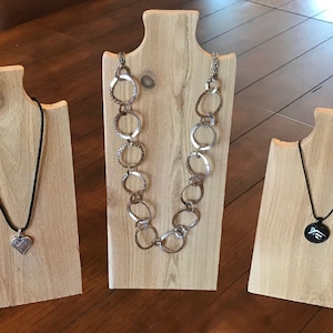May include: Three wooden necklace displays with different necklaces. The display on the left has a silver heart pendant on a black cord. The middle display has a silver chain necklace with large, textured, oval links. The display on the right has a black pendant with a silver design on a black cord.
