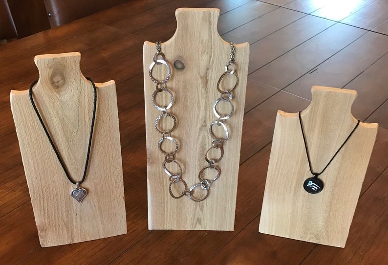May include: Three wooden necklace displays with different necklaces. The display on the left has a silver heart pendant on a black cord. The middle display has a silver chain necklace with large, textured, oval links. The display on the right has a black pendant with a silver design on a black cord.