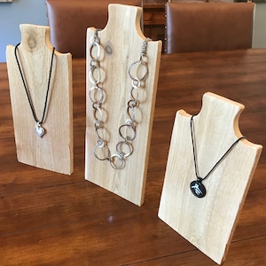 May include: Three wooden necklace display stands with different styles of necklaces. The stands are made of light wood and have a natural finish. The necklaces are made of silver, black cord, and black cord with a silver pendant.