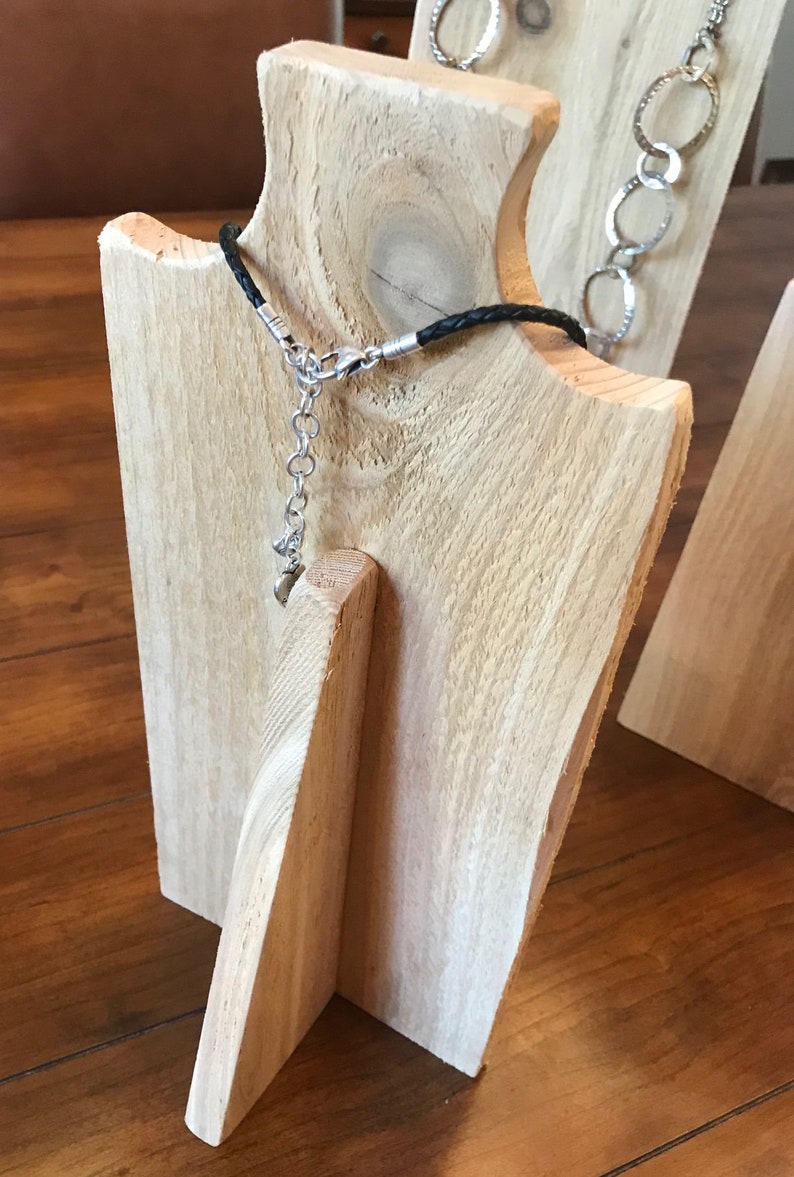May include: A wooden jewelry display stand with a black leather cord necklace. The stand is made of natural wood with a light brown color.