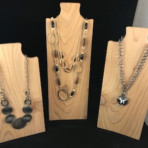 May include: Three wooden necklace displays with different necklaces. The first display has a gold chain with a large pendant made of several round, hammered metal pieces. The second display has a layered necklace with gold, brown, and black beads. The third display has a silver chain with a black and white pendant.