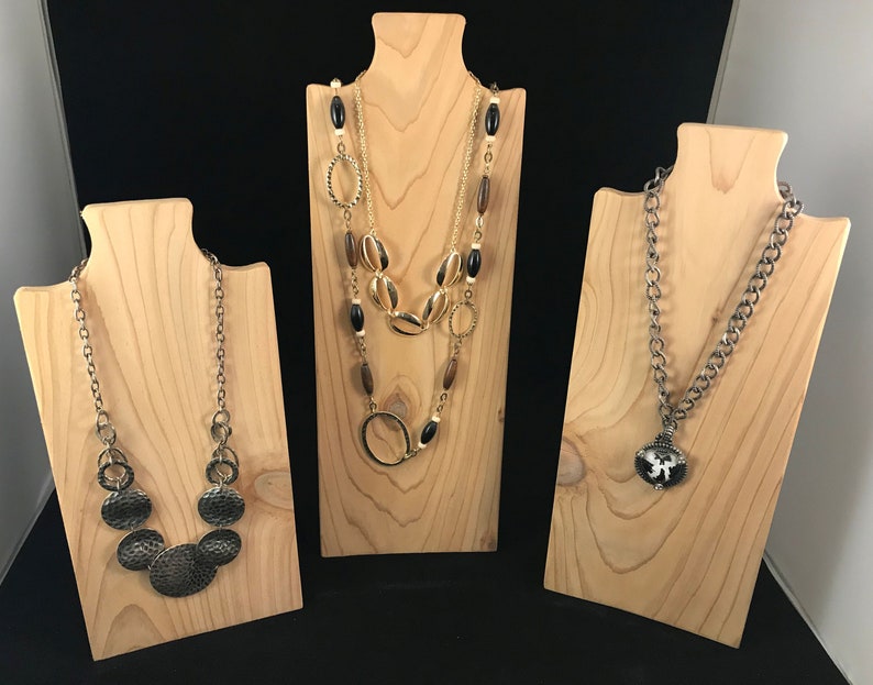 May include: Three wooden necklace displays with different necklaces. The first display has a gold chain with a large pendant made of several round, hammered metal pieces. The second display has a layered necklace with gold, brown, and black beads. The third display has a silver chain with a black and white pendant.