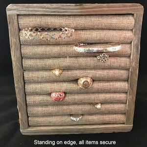May include: A wooden ring holder with a burlap fabric interior. The holder has multiple slots for rings and other jewelry. The holder is standing on its edge, and all of the items are secure.